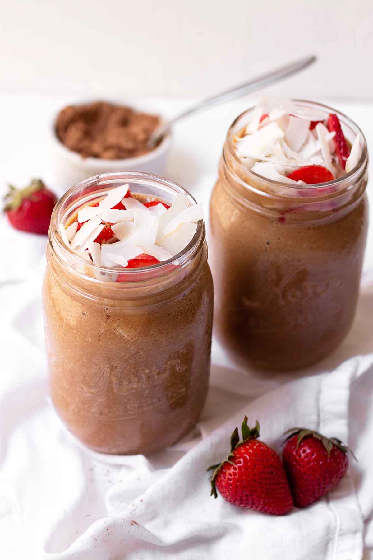 Two glasses of chocolate banana smoothie topped with coconut and strawberries