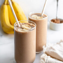 Two glasses of chocolate banana smoothie in front of a bunch of bananas