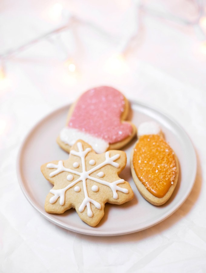 Three Christmas cookies on a plate: an orange light, a pink mitt, and a white snowflake