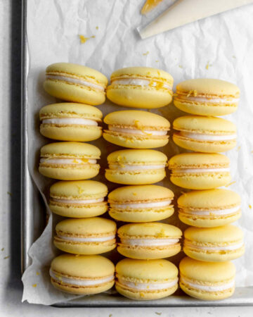 Lemon macarons sitting on a baking sheet next to piping bags with buttercream and lemon curd