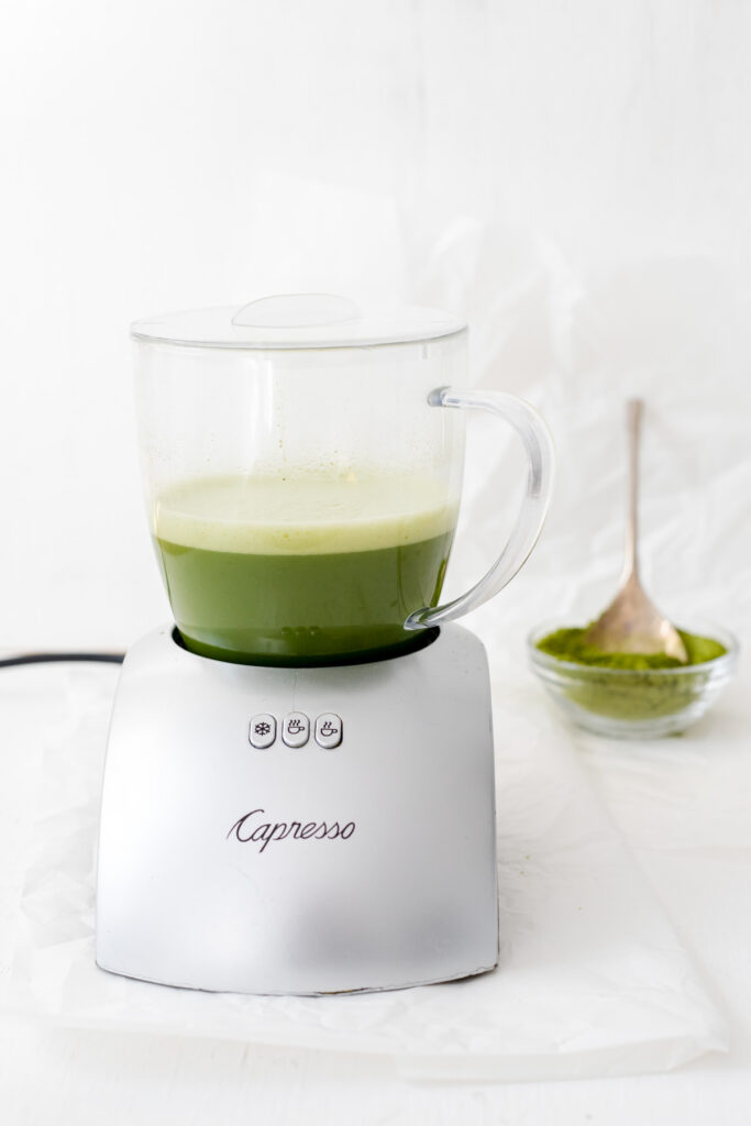 Milk frother with a matcha latte and a bowl of matcha powder with a spoon