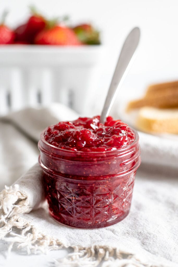 Jar of strawberry jam sitting in front of bread and a basket of strawberries 