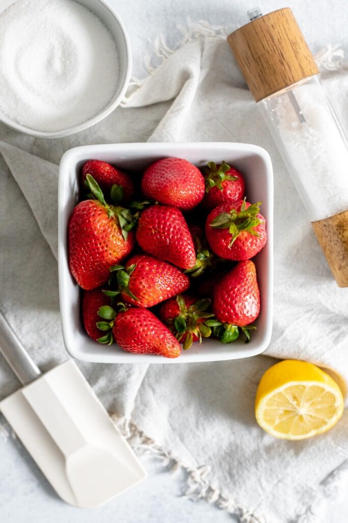 Overhead shot of basket of strawberries, lemon, spatula, salt shaker, and bowl of sugar