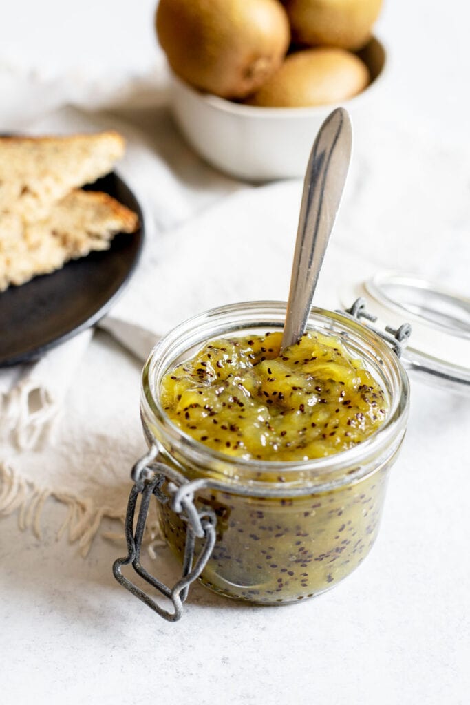 Kiwi jam in a jar with a spoon next to a plate of toast and a bowl of kiwis