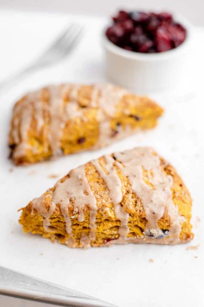 Two pumpkin cranberry scones on a sheet tray in front of a bowl of cranberries