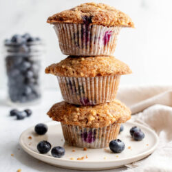 Three blueberry muffins stacked on top of a plate in front of a clear container of blueberries