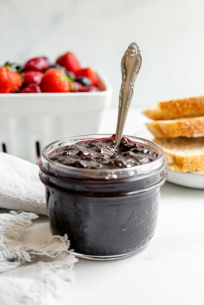Jar of cherry berry jam in front of a basket of fruit and bread