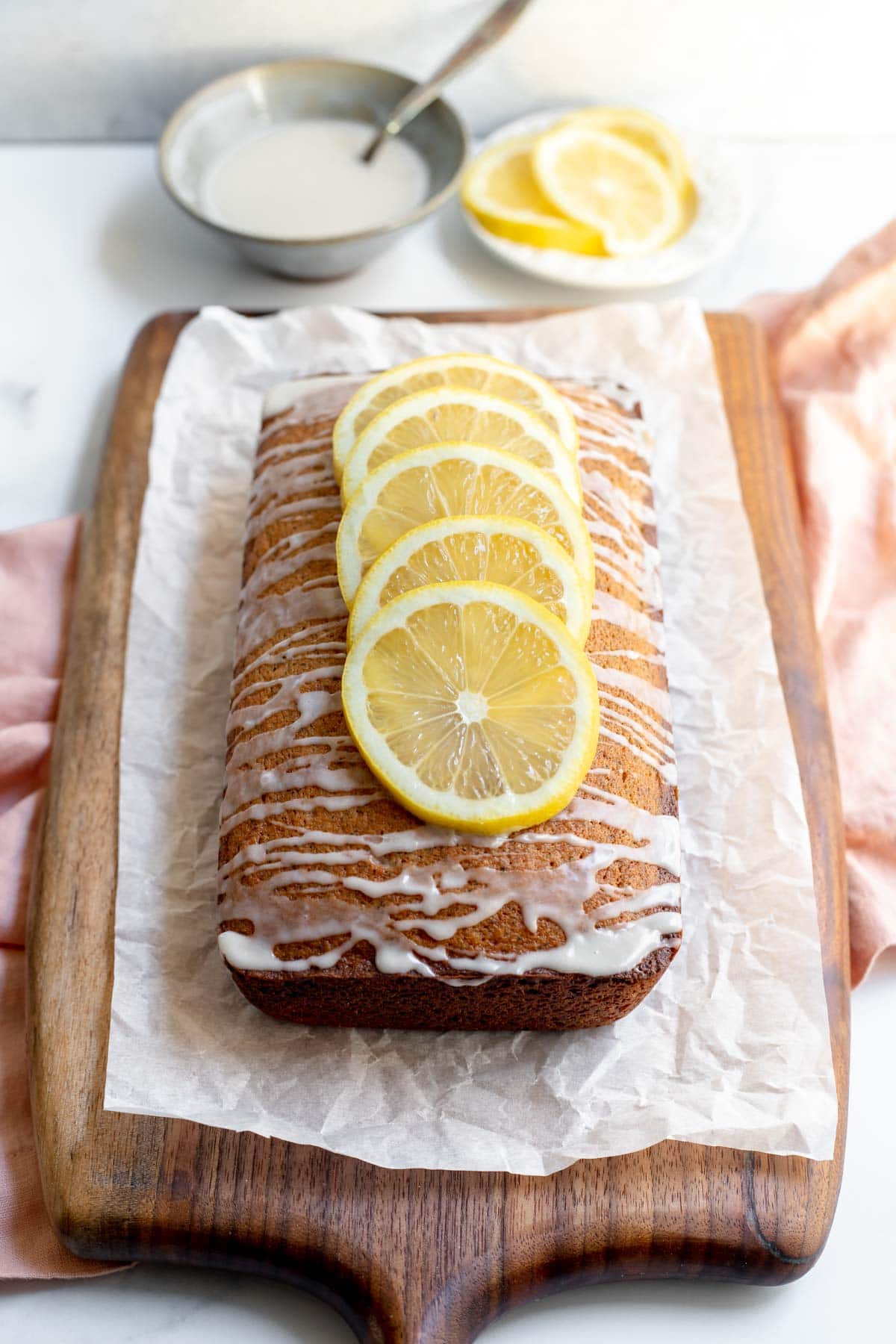 Earl Grey and lemon loaf cake on a cutting board lined with parchment paper in front of a bowl of glaze and a plate of cut lemons