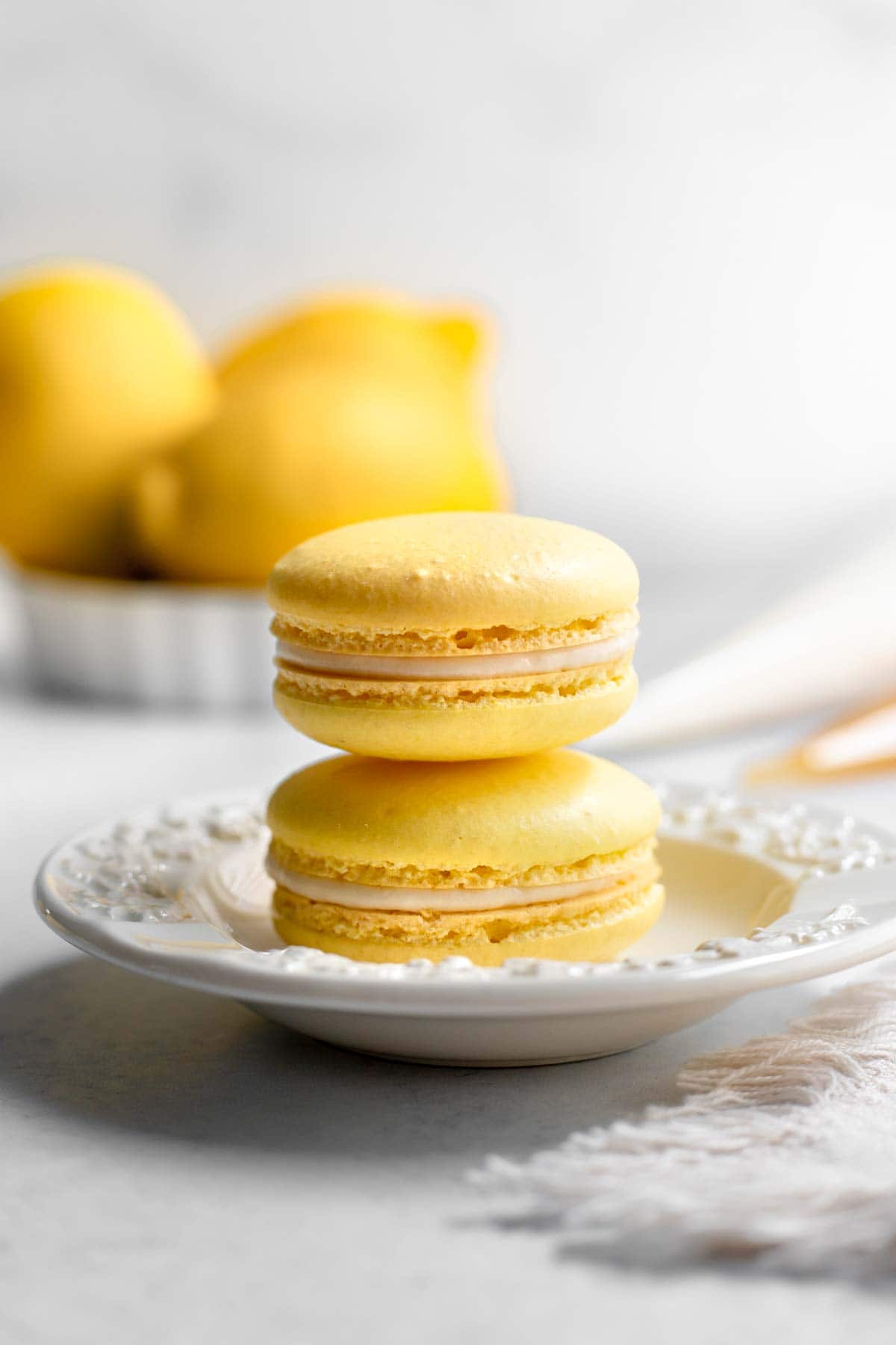 Two lemon macarons stacked on a plate in front of three lemons and piping bags