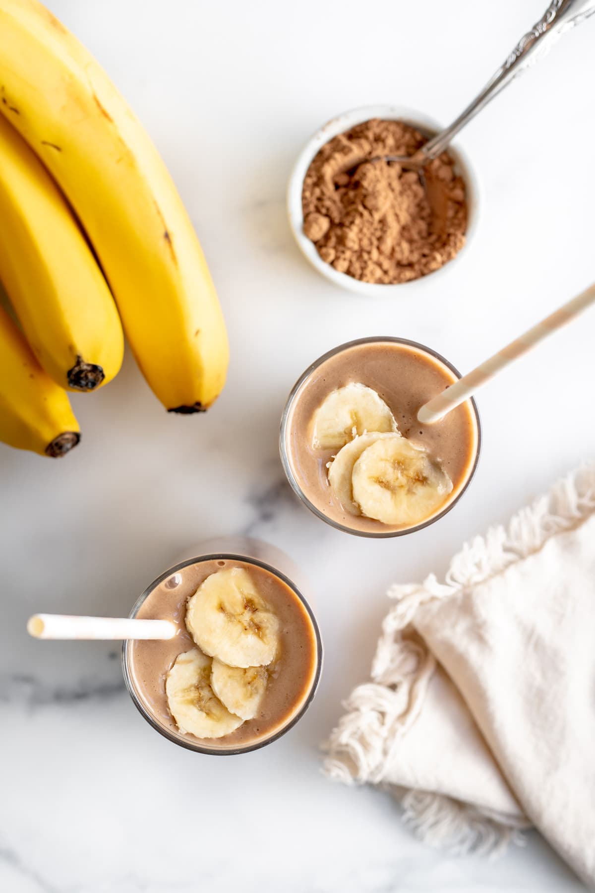 Overhead shot of two glasses of chocolate banana smoothie next to a bunch of bananas