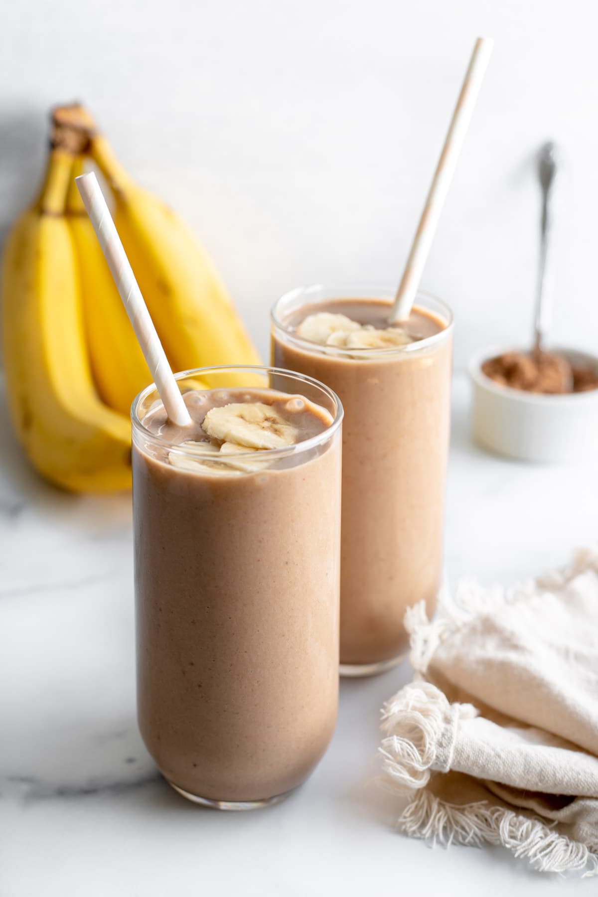 Two glasses of chocolate banana smoothie in front of a bunch of bananas