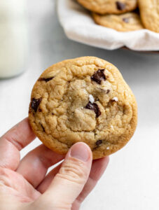 Hand holding cookie in front of a glass of milk and a plate of cookies
