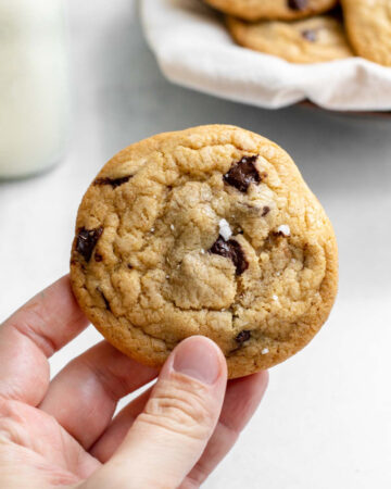 Hand holding cookie in front of a glass of milk and a plate of cookies