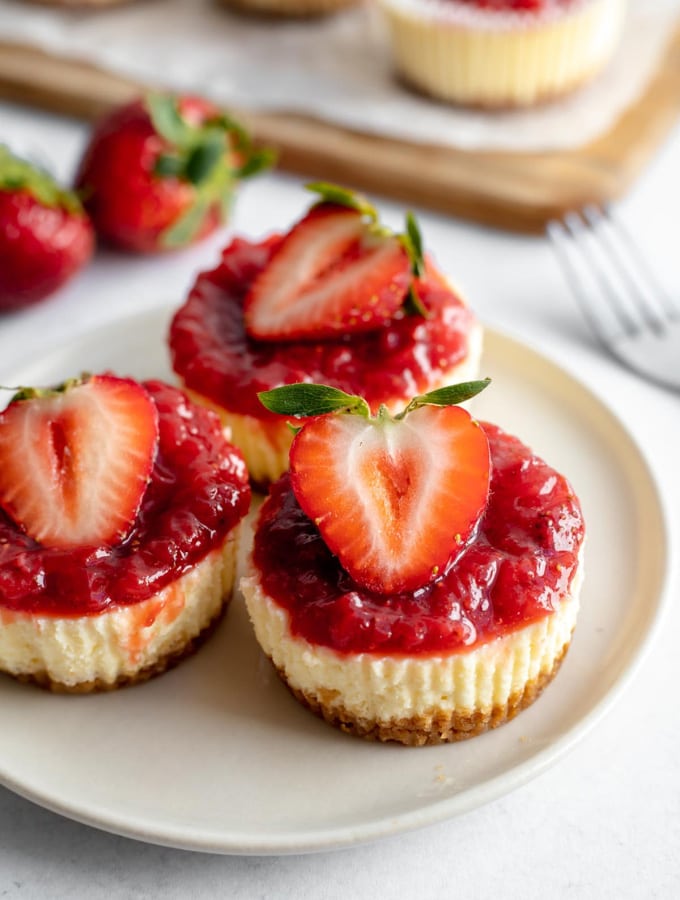 Three mini strawberry cheesecakes on a plate next to a fork and two strawberries