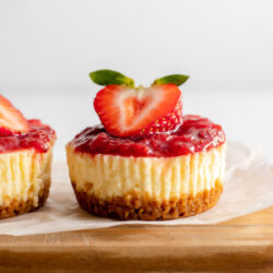 Two mini strawberry cheesecakes on a cutting board with text that reads 'mini strawberry cheesecakes'